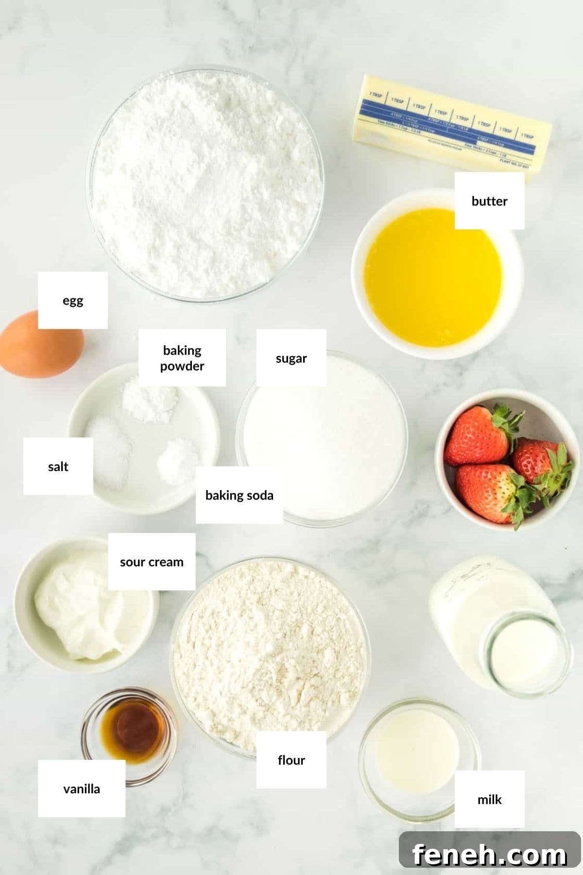 Various baking ingredients for Strawberry Vanilla Cupcakes, including flour, sugar, powdered sugar, butter, egg, milk, sour cream, vanilla, baking powder, baking soda, salt, and fresh strawberries on a white surface.