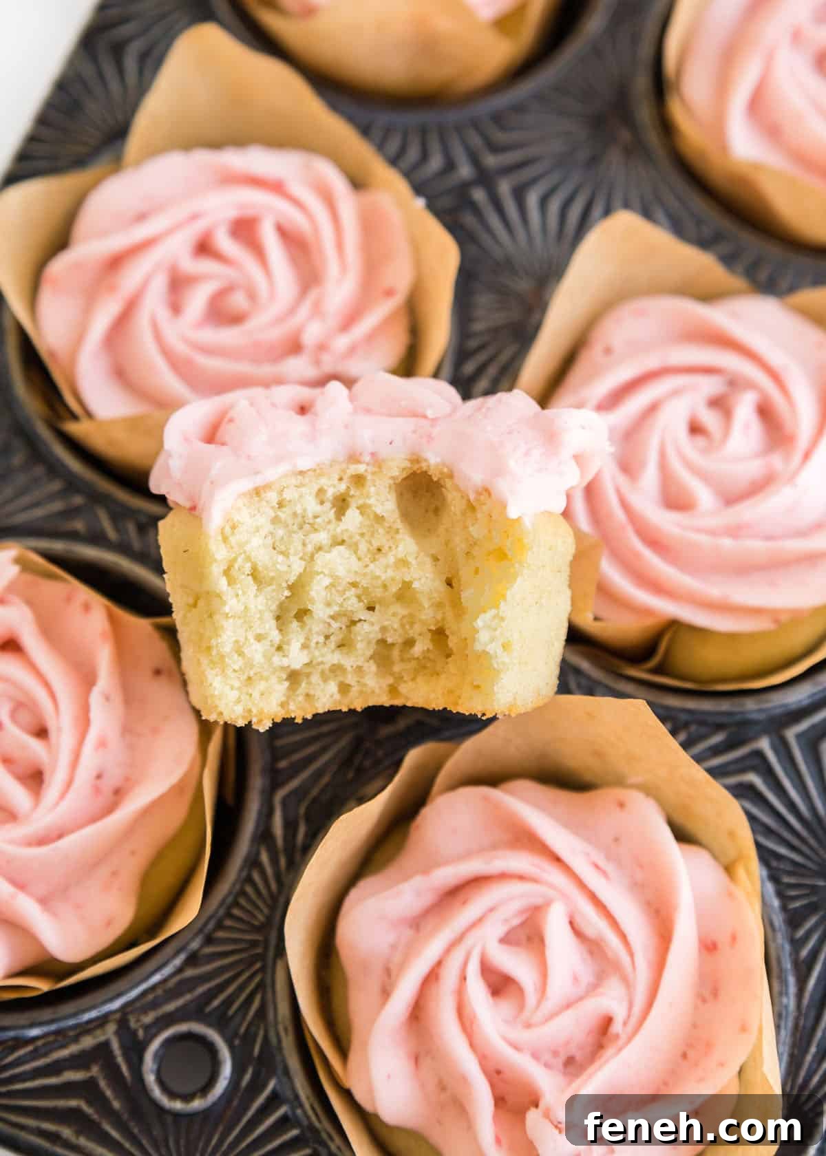 A close up photo of one cupcake with a bite taken out of it placed on top of a muffin tin filled with the strawberry buttercream cupcakes.