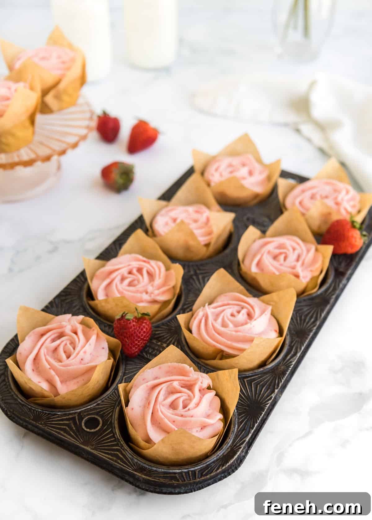 Baked strawberry buttercream cupcakes in a cupcake tin with two cupcakes out of focus on a cake stand in the top left corner accented with whole strawberries and two glasses of milk.