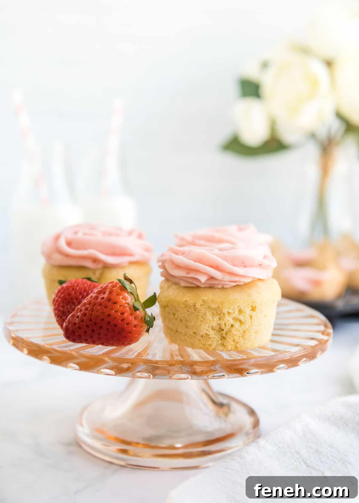 Close-up photo of two cupcakes and two whole strawberries on a small, glass cake stand with two milk jars and a small bouquet blurred in the background.