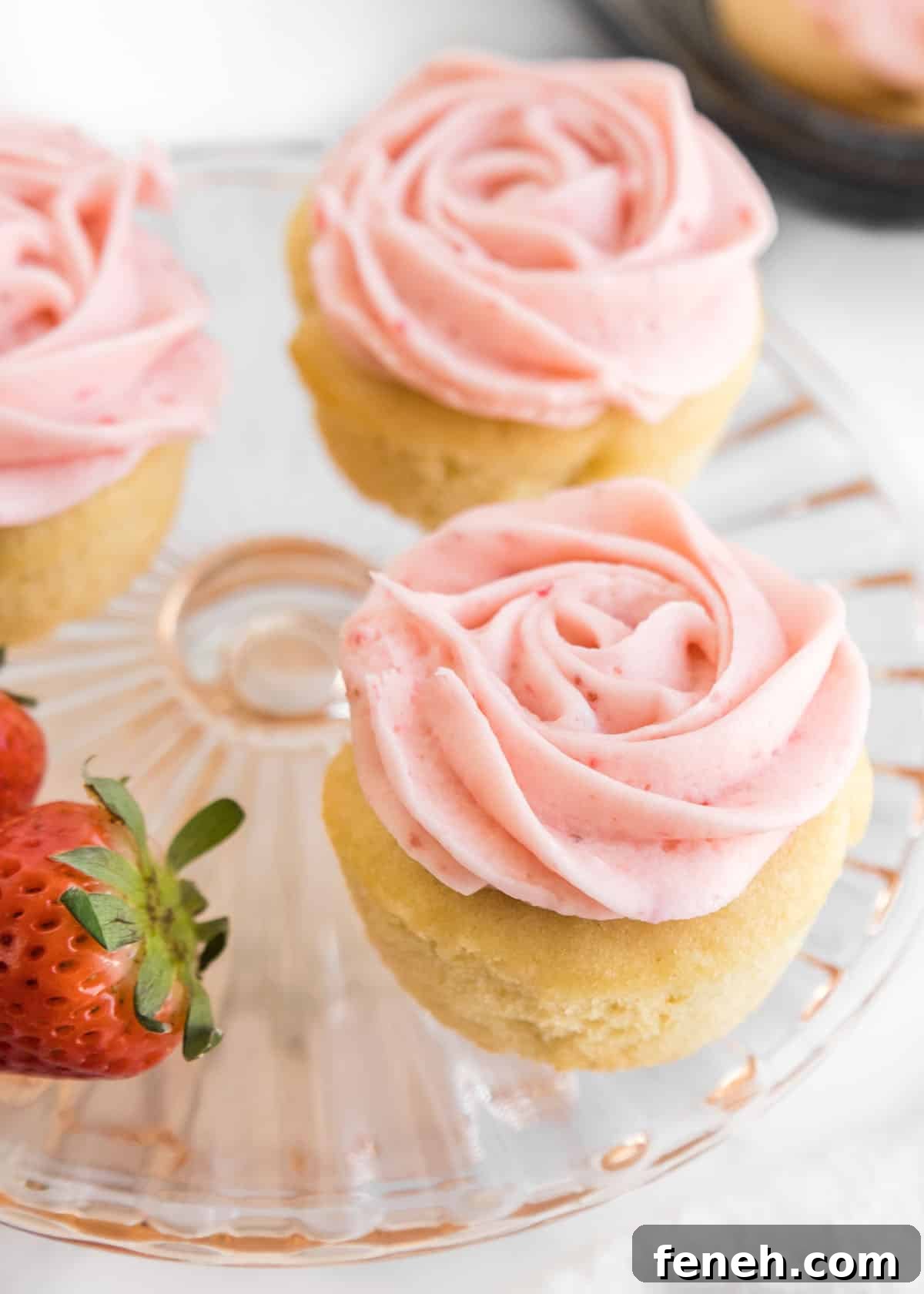 A close-up photo of three cupcakes and two whole strawberries on a small, glass cake stand.