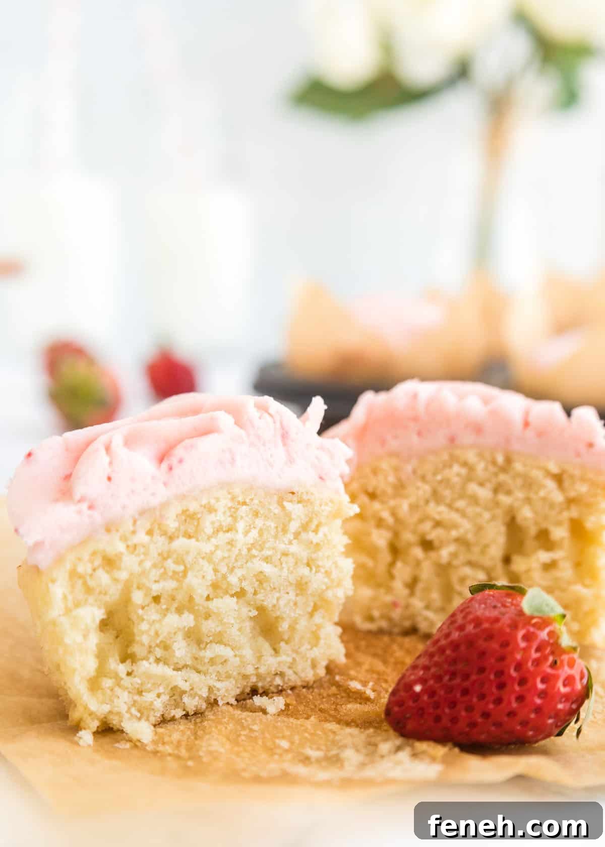 Close-up photo of one cupcake cut in half on top of its parchment liner with one whole strawberry in front of it with extra cupcakes in a muffin tin blurred in the background.