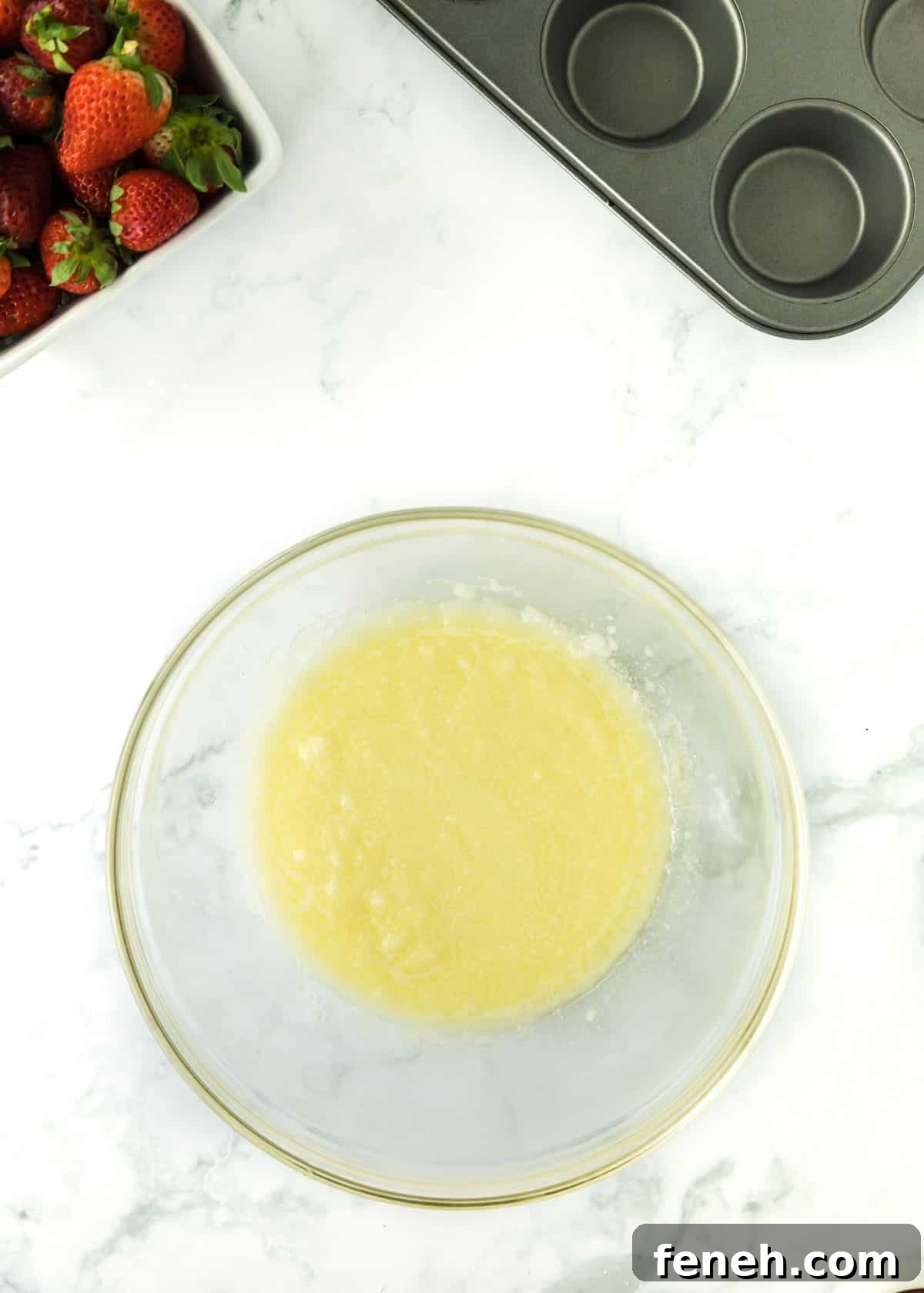 Overhead shot of a glass bowl with melted butter. Upper left and right corners are accented by whole strawberries and an empty muffin tin, respectively.