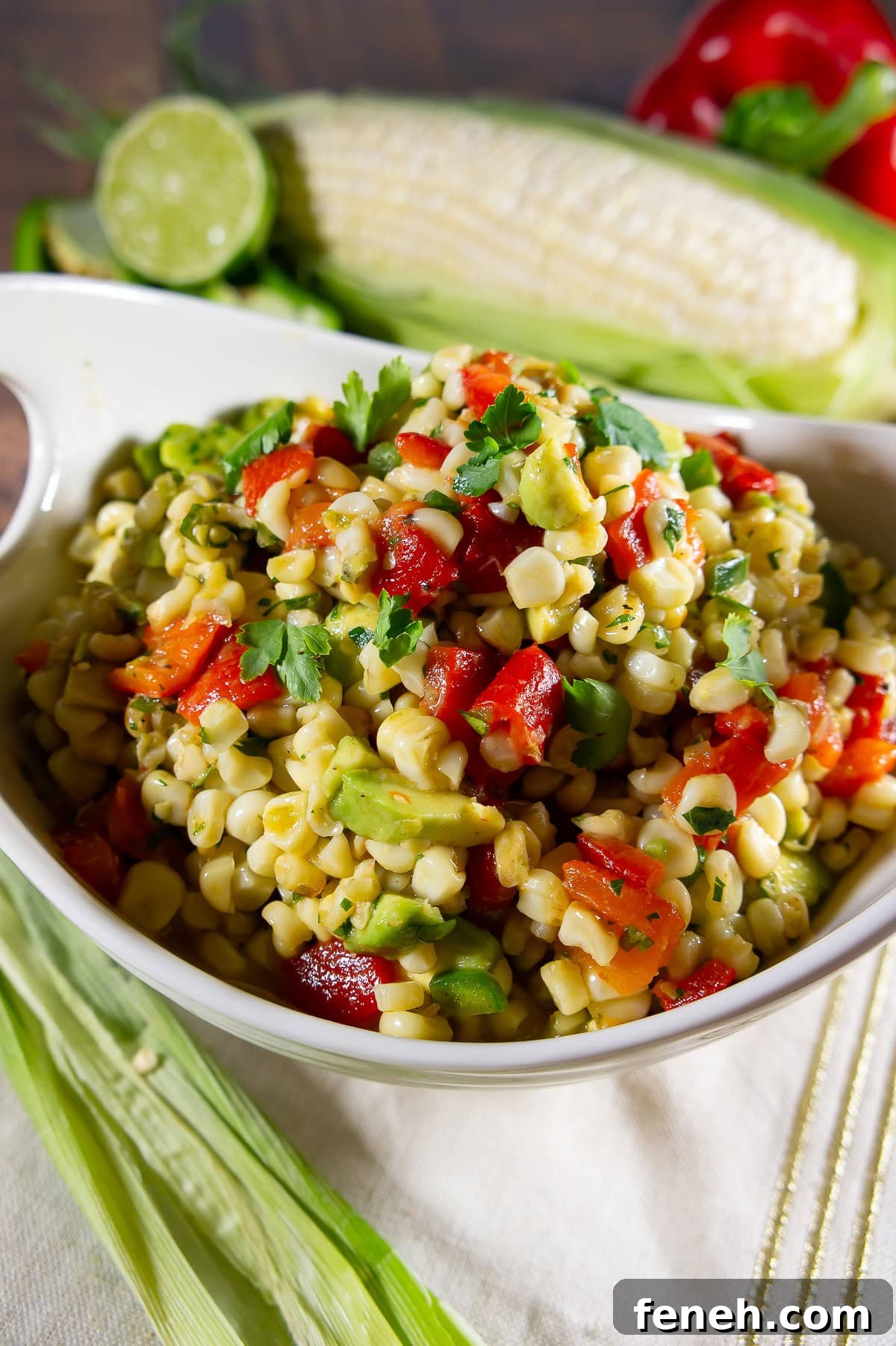 a white bowl with corn avocado salad in it with fresh corn and peppers behind it.