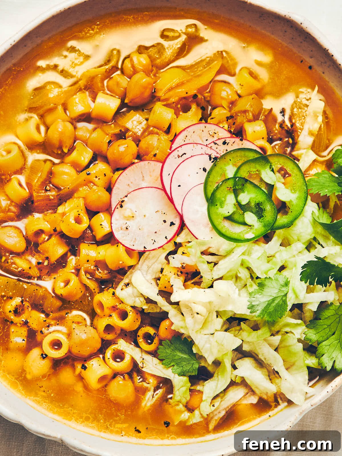An overhead photo of a bowl filled with golden chickpea noodle soup, topped with shredded iceberg lettuce, radishes, and jalapeno peppers