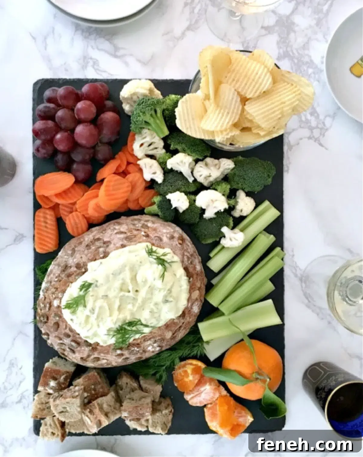 Overhead view of dill pickle dip in a bread bowl, on a black slate snack board with bread cubes, celery sticks, broccoli and cauliflower trees, ruffled potato chips, a bunch of grapes, and sliced carrots.