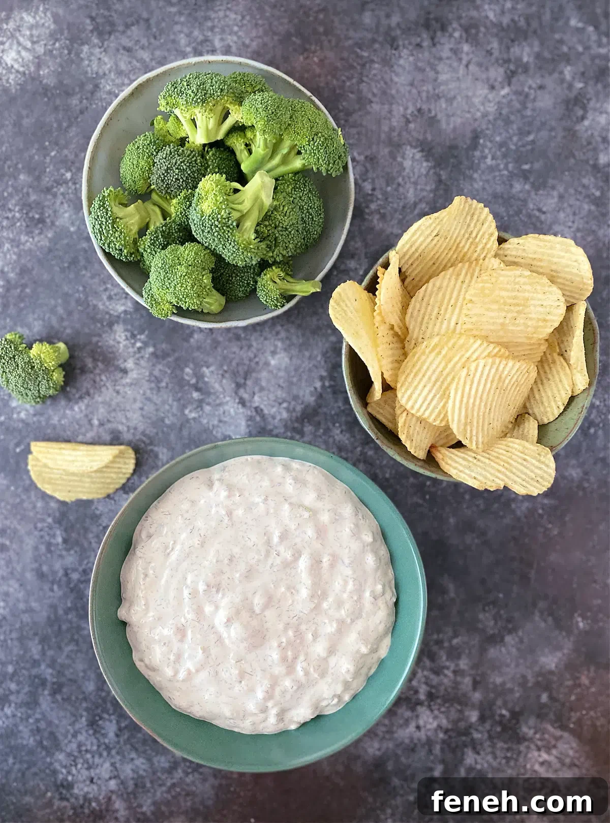 Overhead view of dill pickle dip in a green bowl, with bowls of ruffled potato chips and broccoli trees for dipping.