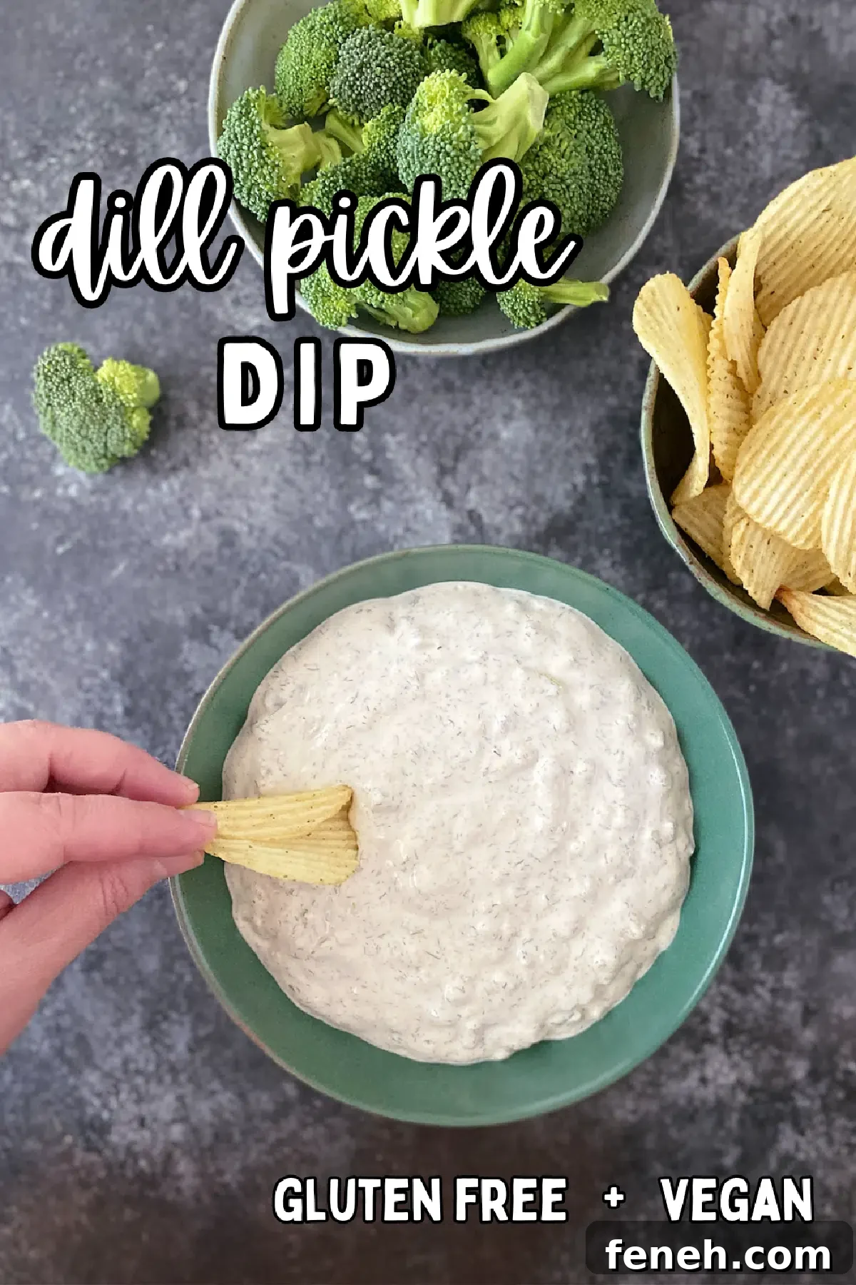Overhead view of dill pickle dip in a green bowl, with a hand dipping a potato chip in the bowl, next to bowls of ruffled potato chips and broccoli trees for dipping.