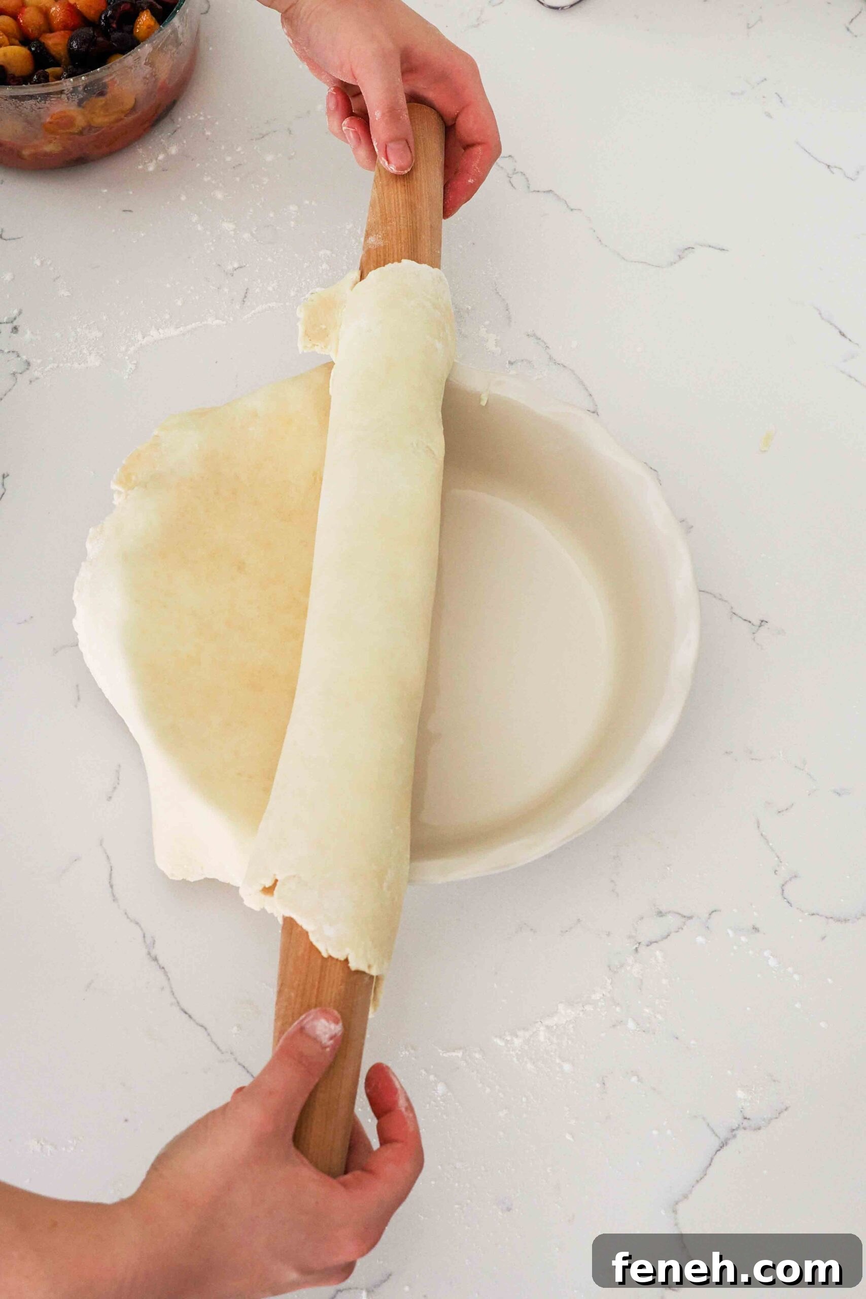 Two hands unroll pie dough from a rolling pin into a pan.