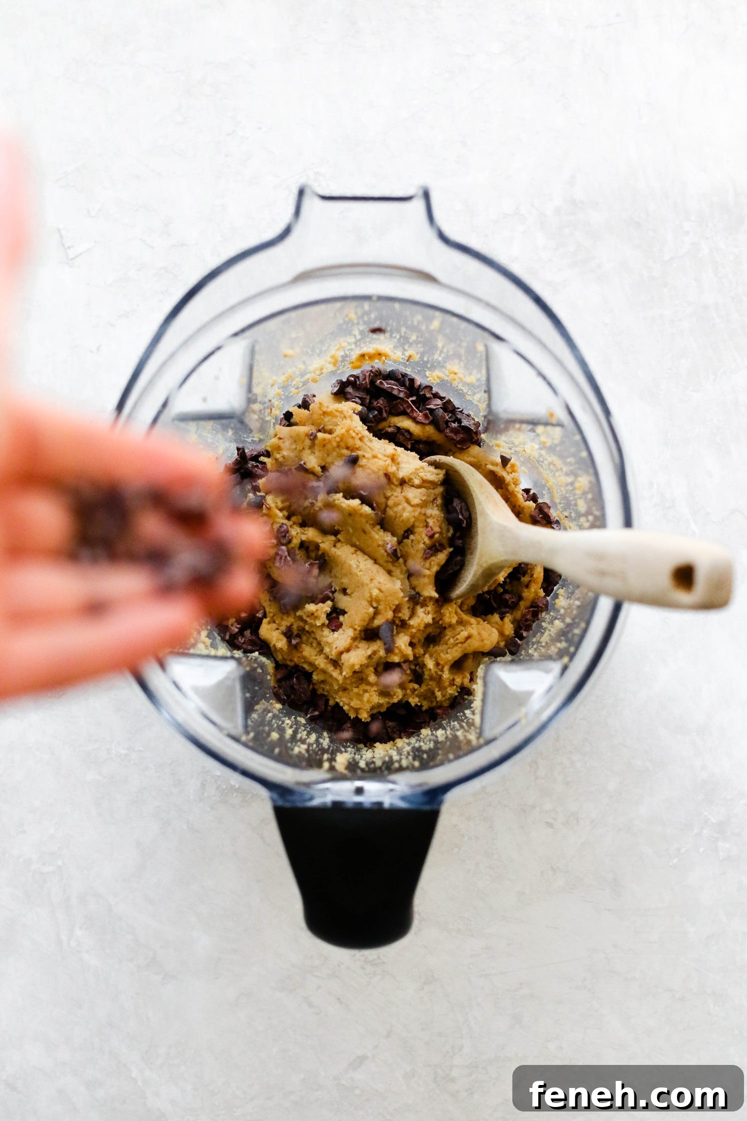 hand adding Cacao nibs to Chickpea Cookie Dough in Vitamix with wooden spoon 