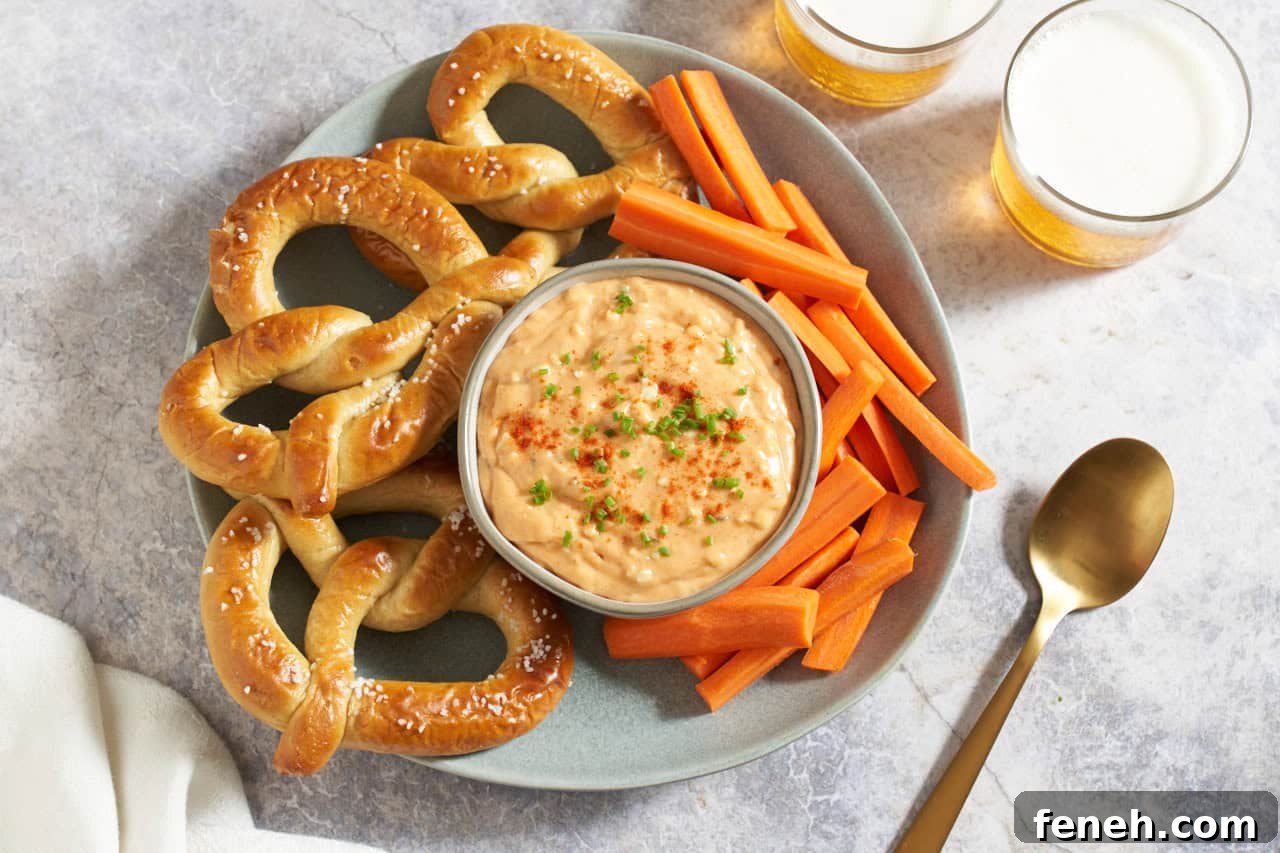 Obatzda german cheese spread in a bowl topped with paprika and chives, surrounded by soft pretzels and carrot sticks, two glasses of beer are in the upper right.