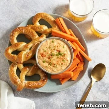 Obatzda german cheese spread in a bowl topped with paprika and chives, surrounded by soft pretzels and carrot sticks, two glasses of beer are in the upper right.