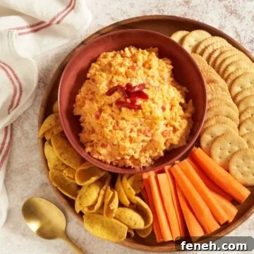 Pimento cheese dip in a red bowl on a round wooden tray, surrounded by butter crackers, carrot sticks, and corn chips.