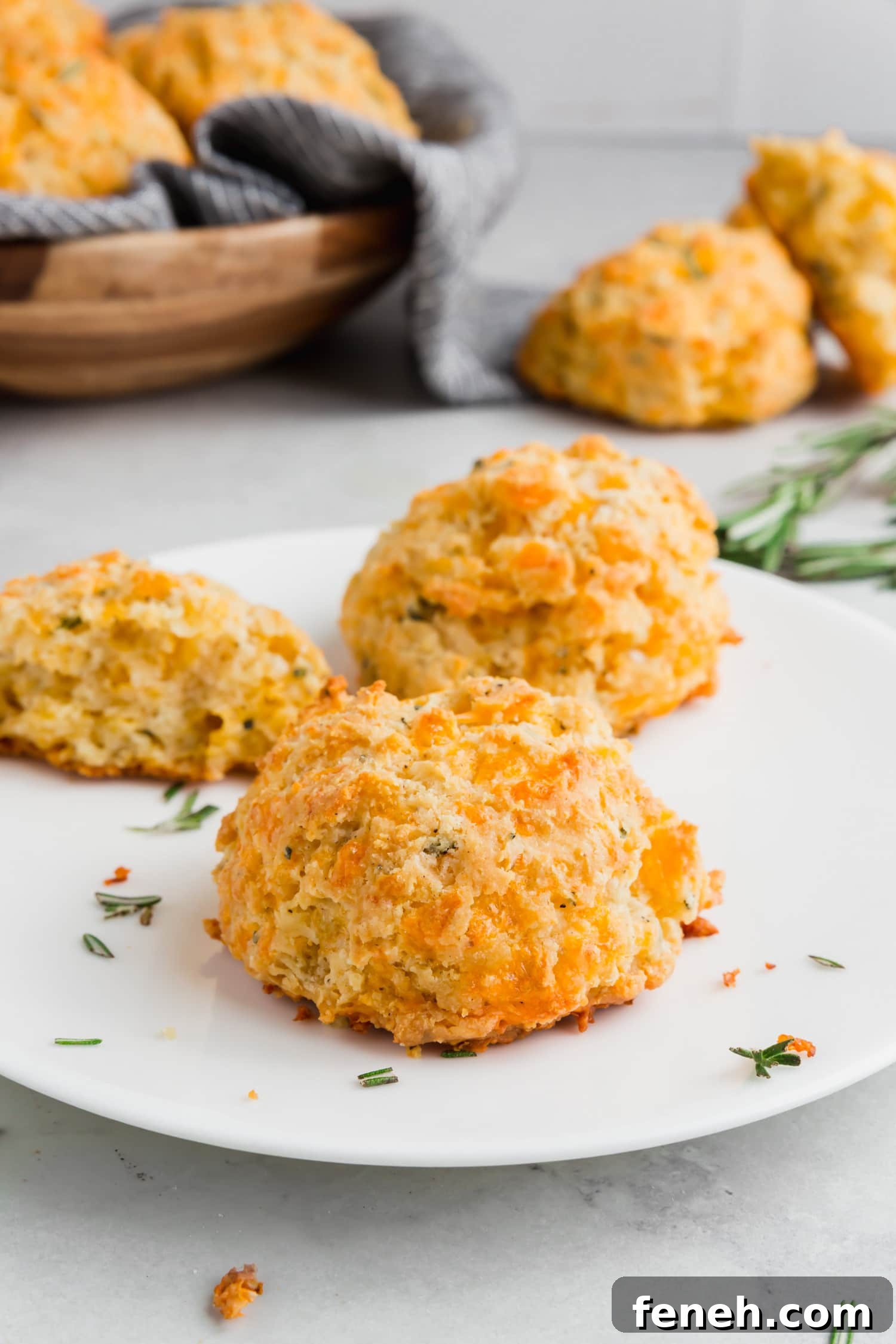 A photo of gluten-free cheese biscuits on a plate with fresh rosemary and a basket of biscuits in the background.