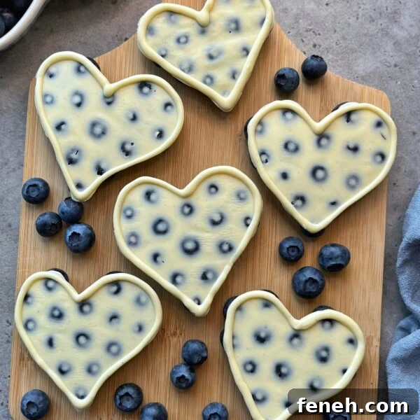 Six heart-shaped cheesecakes with embedded blueberries are arranged on a wooden board, creating delicious bites surrounded by fresh blueberries on a gray surface. Perfect for anyone who loves health-conscious food recipes.