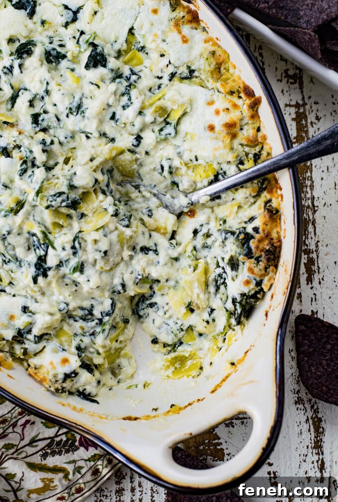 A casserole dish of baked spinach artichoke dip. Blue tortilla chips sit in the corner of the photo along with a green, white and purple napkin.