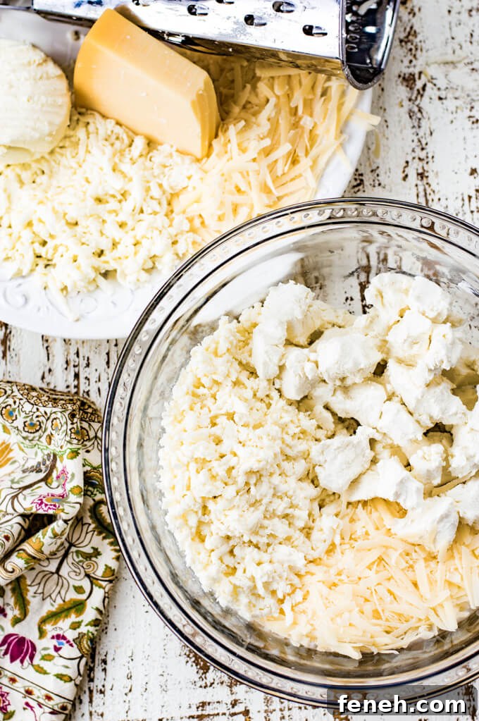 A clear mixing bowl with shredded mozzarella and parmesan and goat cheese. A white bowl sits next to it with a grater and the cheeses. A green, tan and purple napkin sit next to it.