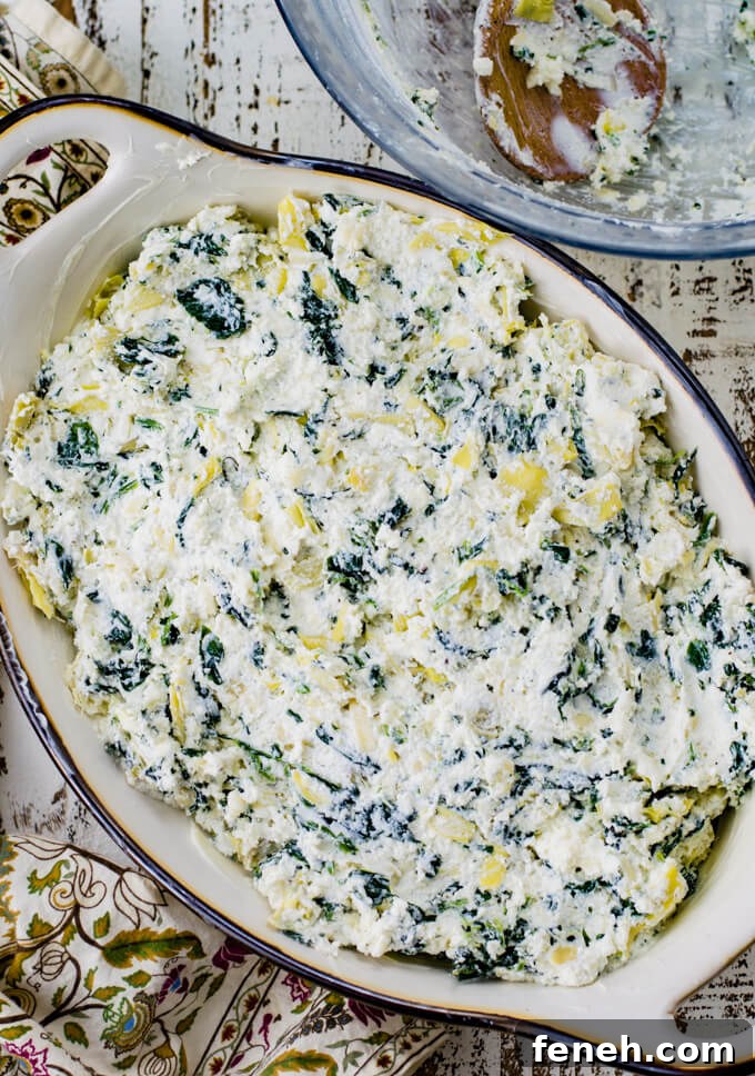 A casserole dish of unbaked spinach artichoke dip. A green, tan and purple napkin sits next to it along with a mixing bowl that had the dip in it. A wooden spoon is in the bowl.