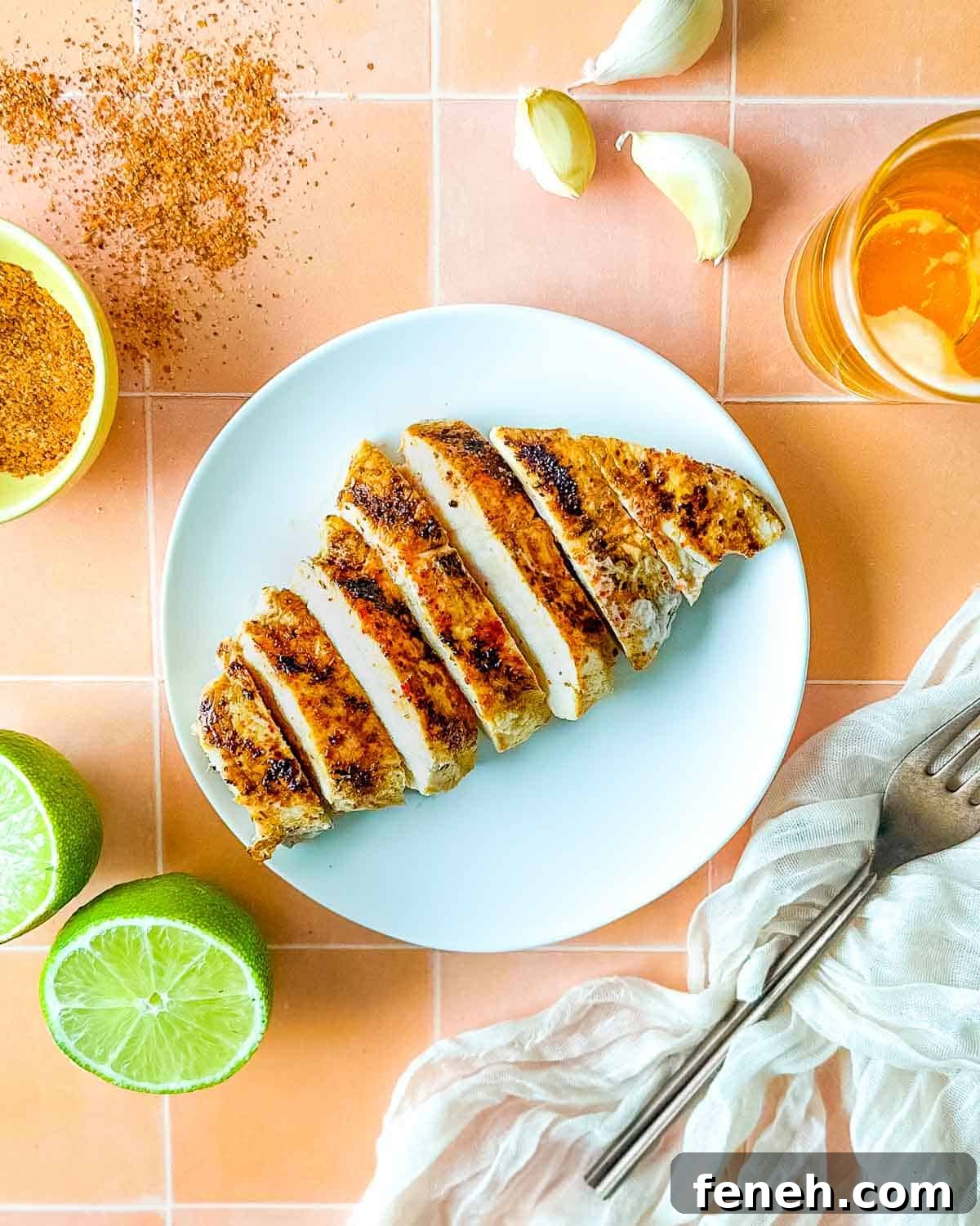 Sliced Tajín Grilled Chicken sits on a white plate surrounded by Tajín seasoning, a sliced lime, a white linen, and a small glass of beer.