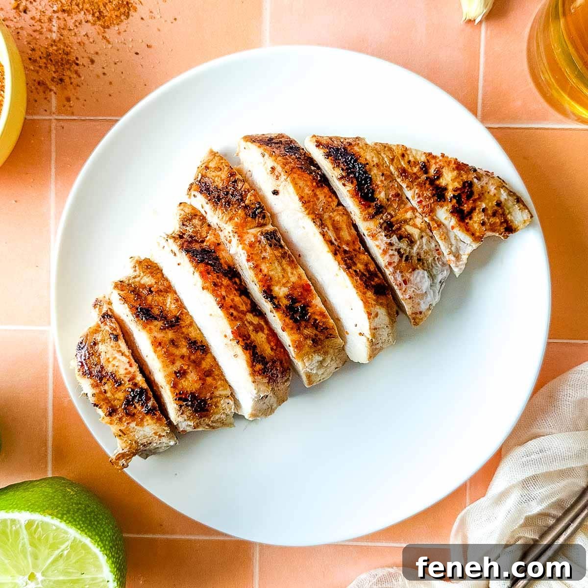 Sliced Tajín Grilled Chicken sits on a white plate surrounded by Tajín seasoning, a sliced lime, a white linen, and a small glass of beer.