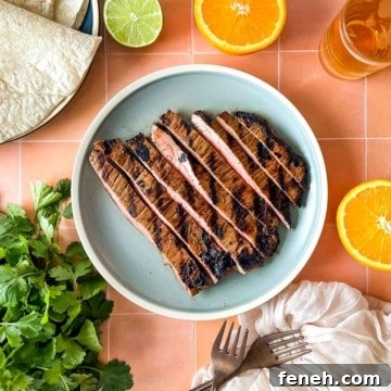 Sliced carne asada sits on a light blue plate surrounded by sliced oranges, limes, a bunch of cilantro, tortillas, and a glass of beer.