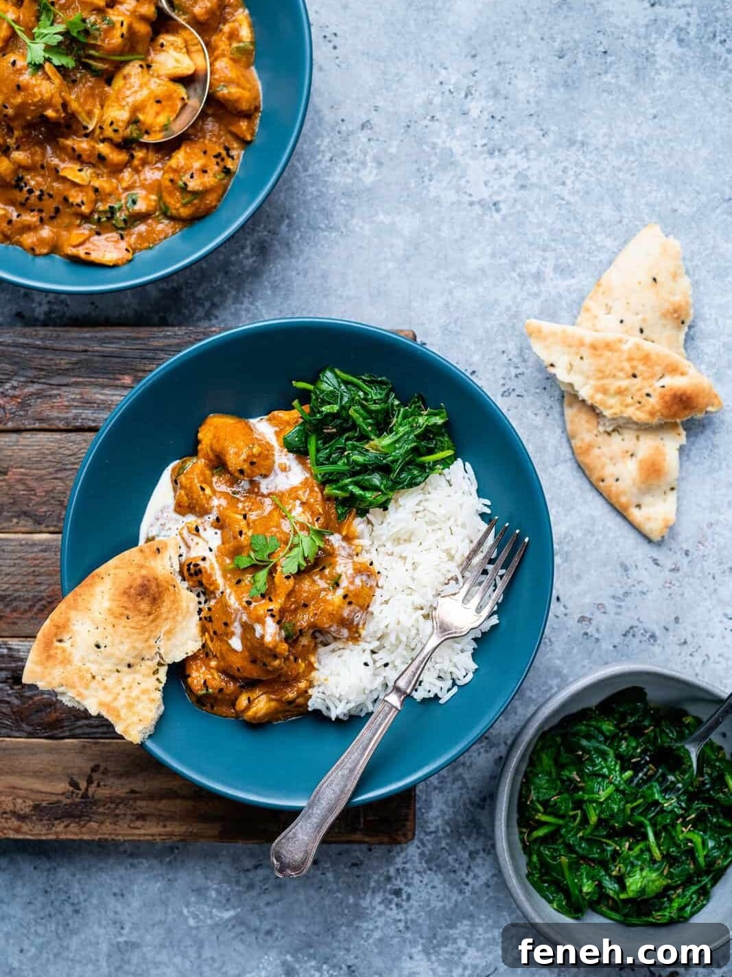 A blue bowl filled with rice, creamy easy curry recipe, sautéed spinach, and naan bread, with a fork on the side. Another bowl, naan pieces, and a small bowl of spinach are nearby on a gray background.