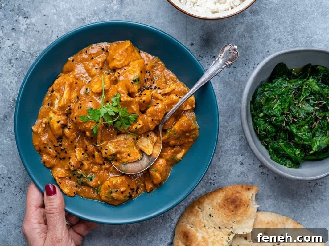 A hand holds a blue bowl of creamy paneer curry with a spoon, garnished with cilantro. Perfect as an easy curry recipe, it’s served alongside white rice, sautéed greens, and naan on a gray textured surface.