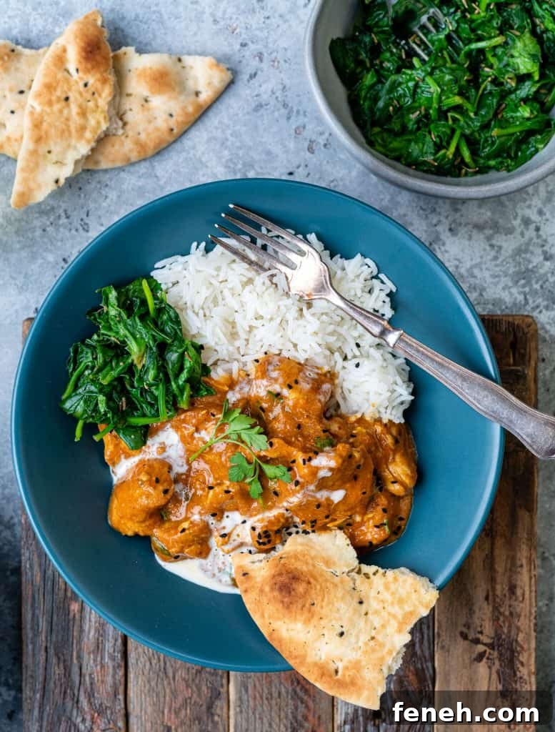 A blue bowl with white rice, creamy orange slow cooker chicken curry, sautéed greens, and naan bread, garnished with herbs; a fork rests on top. Nearby, extra naan and greens complete this inviting and easy curry recipe on a rustic wooden board.