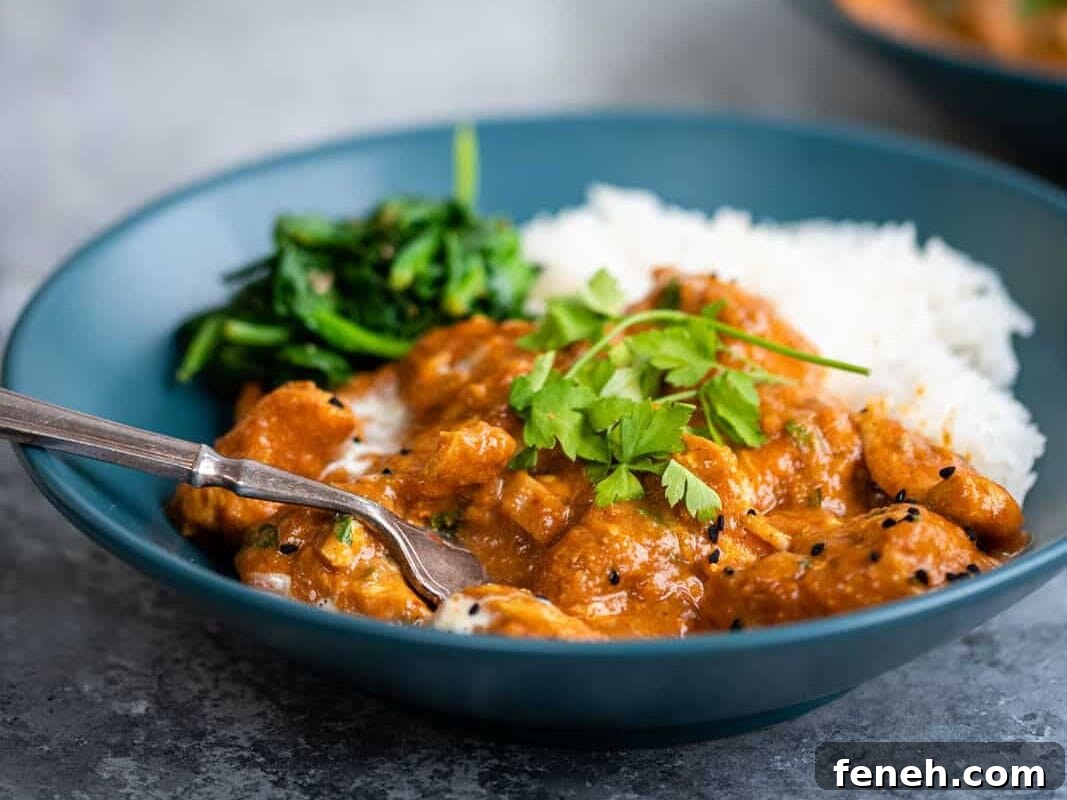 A blue bowl filled with white rice, creamy orange Slow Cooker Chicken Curry, sautéed greens, and cilantro garnish. A fork rests along the bowl’s edge; another bowl and glass are blurred in the background.