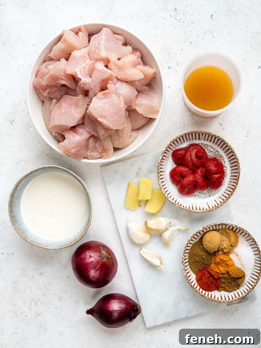 Top-down view of Slow Cooker Chicken Curry ingredients: raw diced chicken, broth, cream, spices, tomato paste, ginger, garlic cloves, and whole red onions—everything you need for an easy curry recipe all laid out on a white surface.