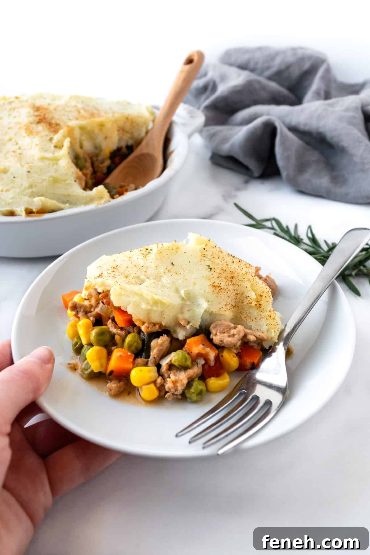 A hand holding a white plate with a piece of shepherds pie on it with a fork.