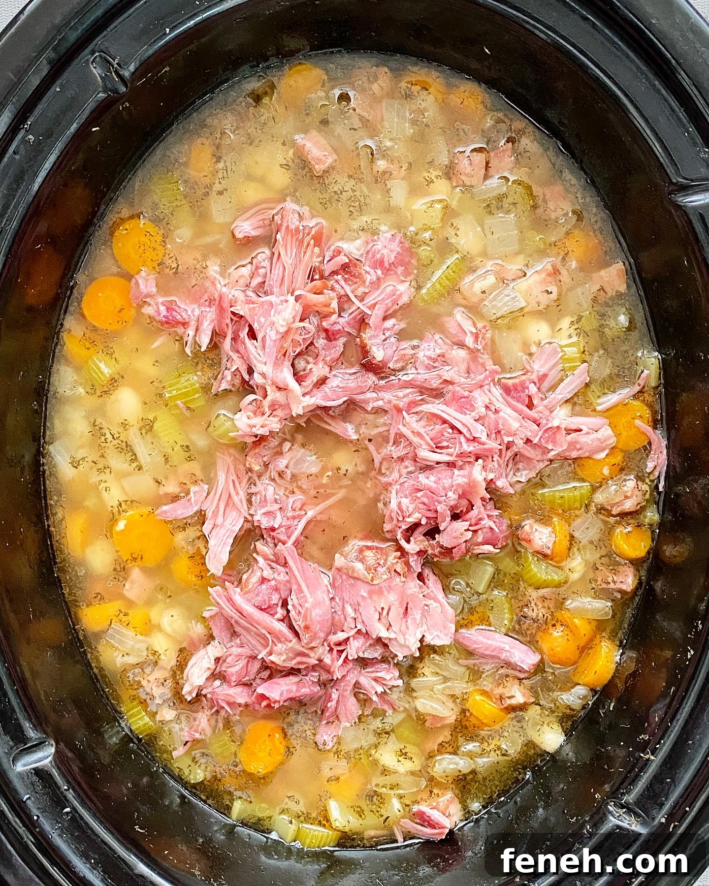 overhead shot of cooked crockpot ham and bean soup in a black slow cooker with shredded ham hock meat on top