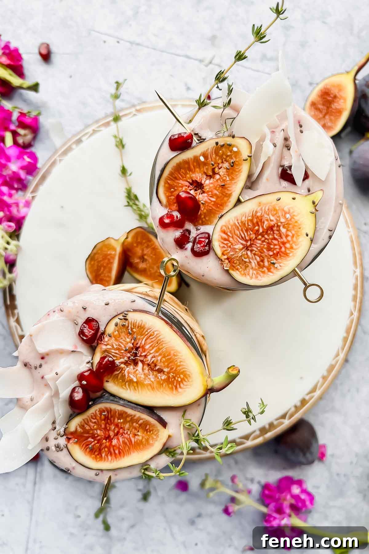 overhead shot of two fig smoothies garnished with fresh fig halves, pomegranate seeds, and coconut flakes on white serving tray with flowers and greenery surrounding.