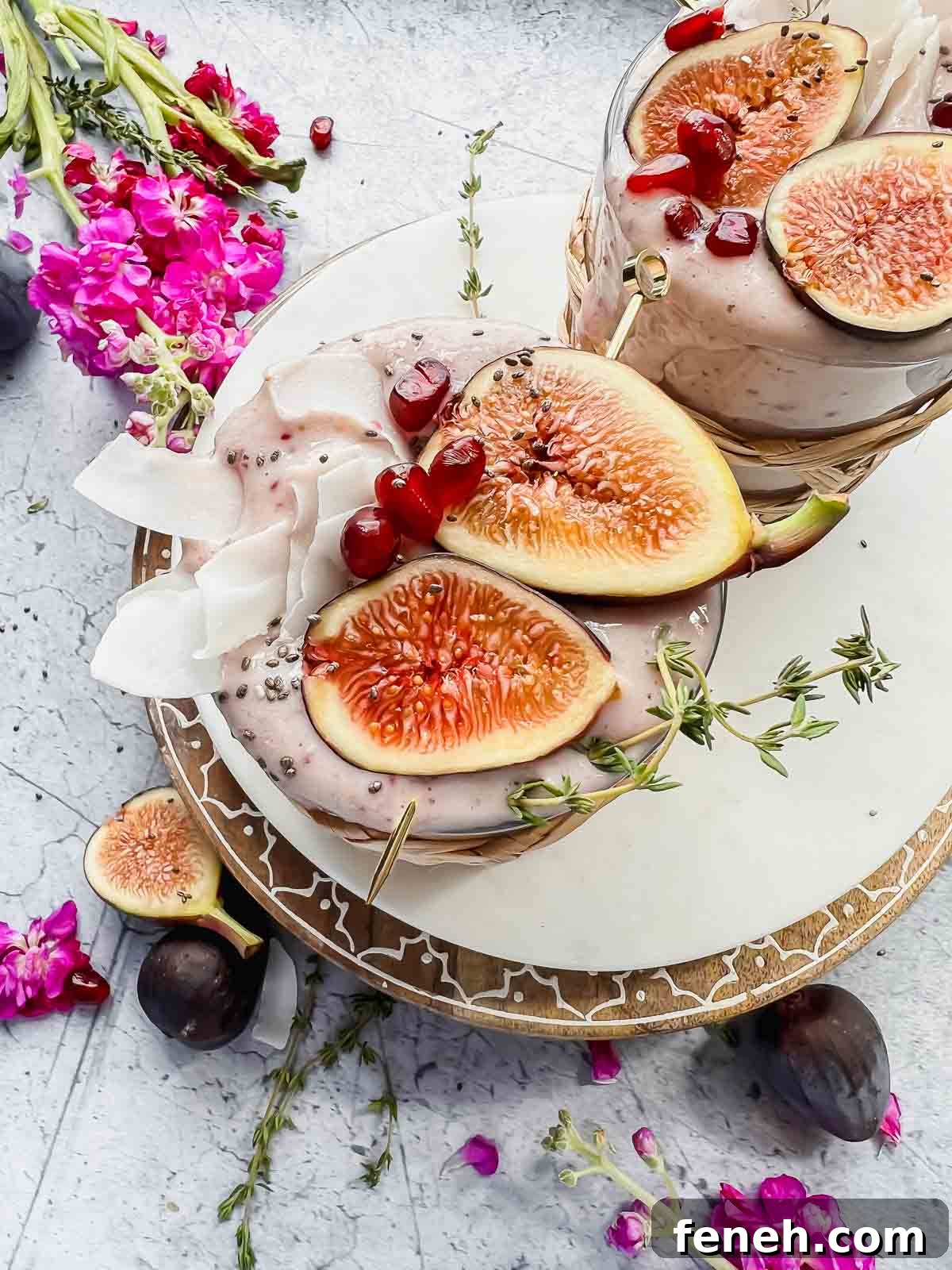 fig smoothie garnished with fresh fig halves, pomegranate seeds, and coconut flakes in glass with wicker sleeve on white tray with additional fig halves surrounding and vase of flowers in background.