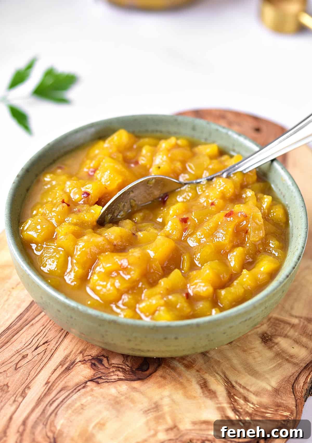 Wooden board with green bowl containing mango chutney. A silver spoon, white background.